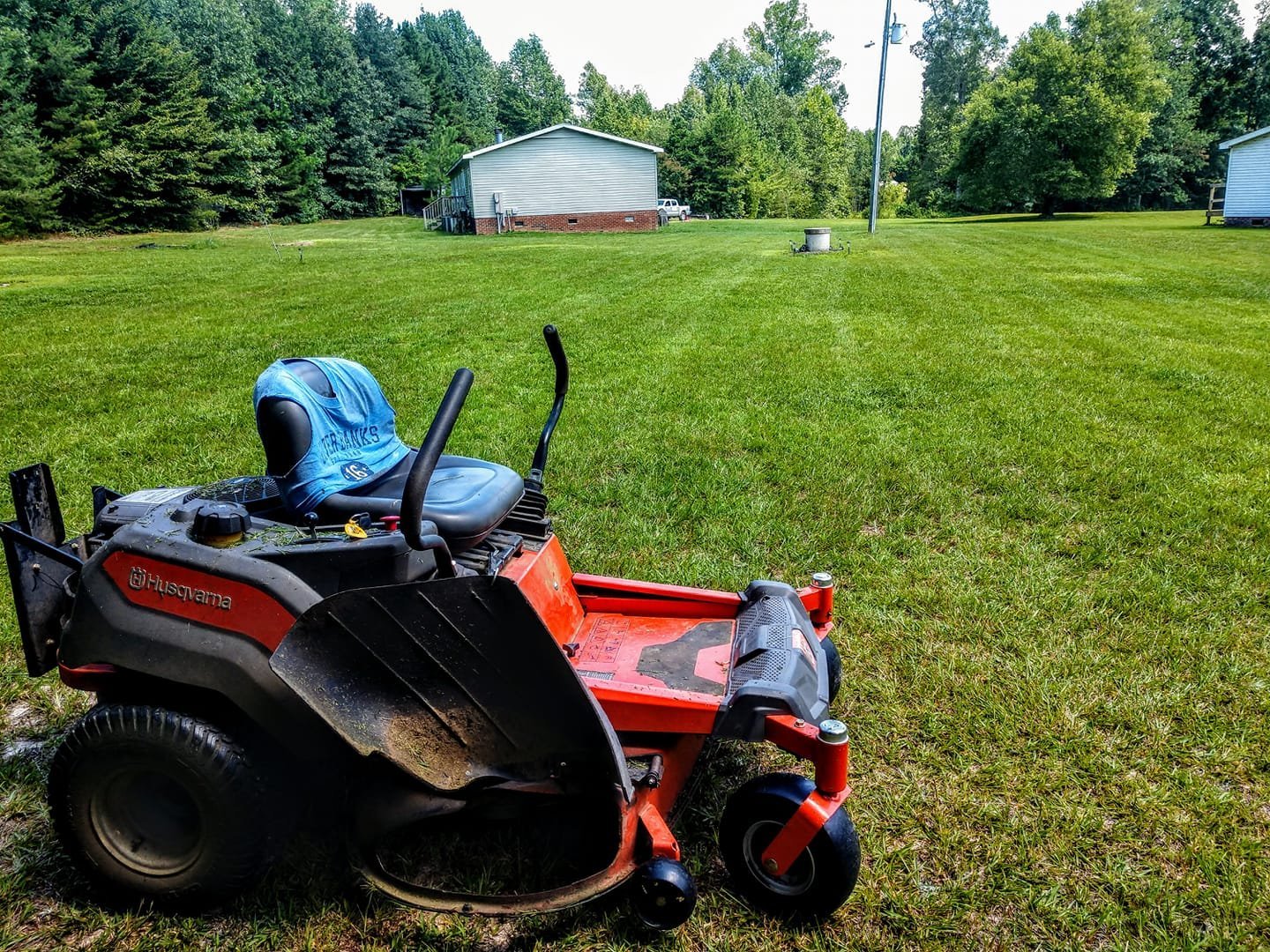 Red and black riding lawn mower with blue canopy in large green field with tree-lined background and farm buildings