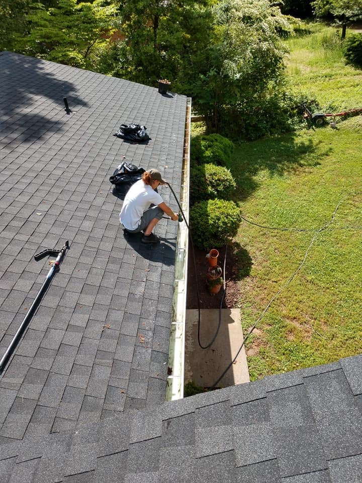 Worker in white shirt on gray shingled roof performing maintenance or inspection with tools and equipment, overlooking green yard